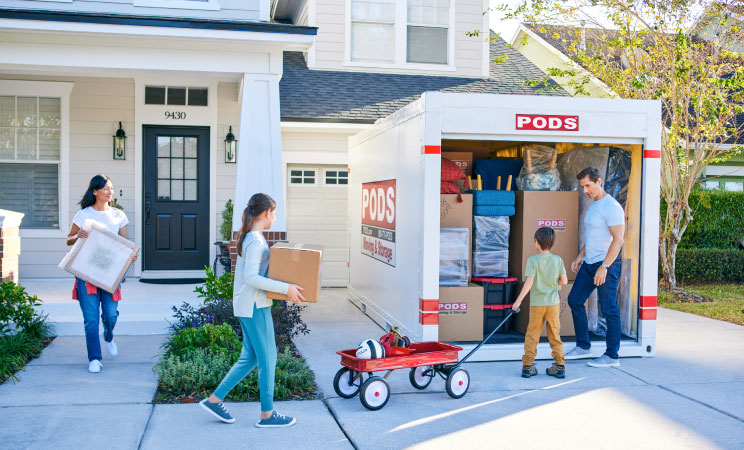 A family of four carries a few final items to the PODS portable moving container sitting in their driveway. The container is nearly full, having been expertly loaded with boxes and furniture.